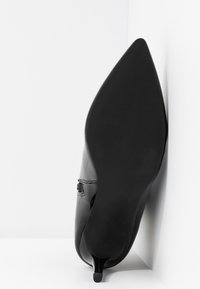 Chaussure à talon kitten en cuir verni noir à bout pointu, positionnée semelle vers le haut contre un angle blanc entre un mur et le sol.