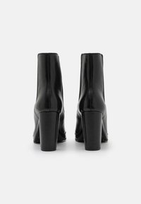 Botas de tornozelo em couro preto com acabamento brilhante, saltos quadrados e costura lateral. O design apresenta uma silhueta elegante e uma textura suave.