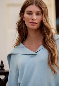 Gaiši zils džemperis ar V veida izgriezumu un platu apkakli, ar mīkstu tekstūru un vaļīgu piegriezumu, kas brīvi krīt pār pleciem.