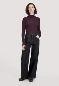 Frau mit kurzen dunklen Haaren, die einen figurschmeichelnden dunkelvioletten Rollkragenpullover und weite schwarze Jeans trägt, mit den Händen in den Taschen, stehend vor einem hellen Hintergrund.