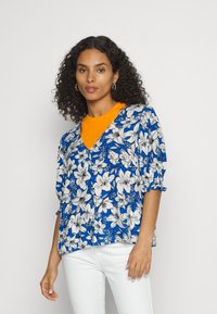 Blusa azul floral com padrões de flores brancas, com mangas curtas e design em V, combinada com uma t-shirt laranja brilhante por baixo.