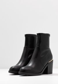 Bottines noires en cuir avec un talon bloc haut et un accent doré sur le talon. Texture lisse et détails cousus autour du haut.