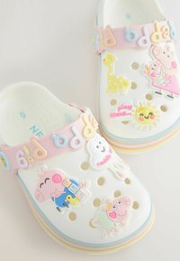 Farverige børnetræsko lavet af hvidt syntetisk materiale, med legesyge tegneserieklistermærker, en lyserød rem og ventilationshuller.