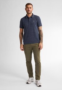 Camisa polo a rayas azul marino con cuello y logo, combinada con pantalones verde oliva y zapatillas blancas. La tela tiene textura.