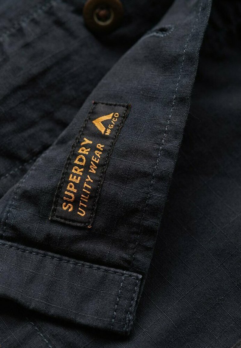 Camisa de tela azul oscuro con superficie texturizada y cierre de botones. Etiqueta "Superdry Utility Wear" en naranja, adjunta al cuello.