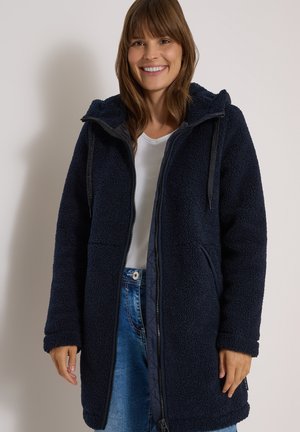 Femme souriante portant un manteau en polaire bleu marine avec capuche, une chemise blanche et un jean bleu sur un fond clair uni.