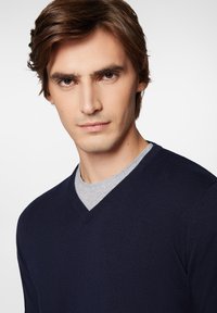 Granatowy sweter z dekoltem w serek o prążkowanej fakturze, noszony na szarym podkoszulku z okrągłym dekoltem, ukazujący zbliżenie rysów twarzy na neutralnym tle.