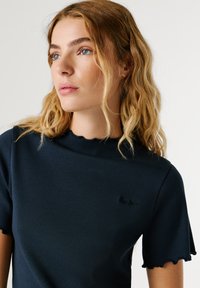 Μπλε σκούρο μπλουζάκι με κοντά μανίκια και ριμπ ύφανση, με σκουληκόστομο ντεκολτέ και τελείωμα, διαθέτοντας ένα μικρό μαύρο λογότυπο στο αριστερό στήθος.