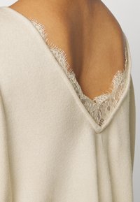 Lichtbeige gebreide top met een v-hals en delicate kantafwerking bij de halslijn. Zachte textuur, met ruwe randdetails langs het kant.