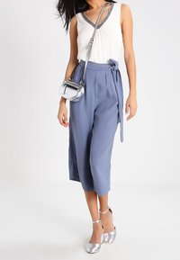 Kvinnaklädd i ärmlös vit blus, högmidjade blå cropped-byxor med sidofäste, silverfärgad axelremsväska och silverfärgade klackar med ankelrem.