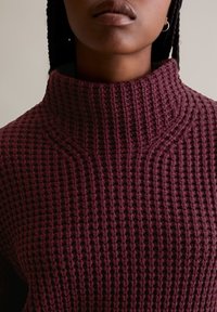 Burgunderfarbener Pullover mit hohem Kragen, der ein strukturiertes Waffelstrickdesign und ausgeprägte Nähte aufweist.