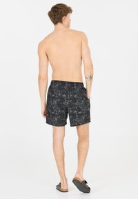 Zwarte zwemshorts met een tropisch patroon, elastische tailleband en zijzakken. Gestyled met instapsandalen en een naakt achteraanzicht.