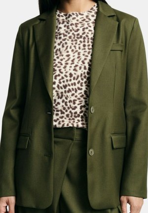 Mujer con un traje verde oliva con una chaqueta de dos botones y pantalones a juego, combinados con una camiseta estampada de leopardo blanca y marrón.