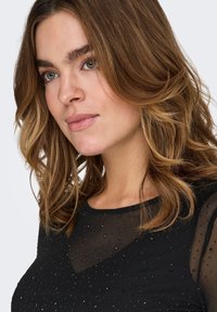 Μαύρο τοπ με διαφανές ύφασμα, διακοσμημένο με μικρές χάντρες. Διαθέτει στρογγυλή λαιμόκοψη και χαλαρά, κυματιστά μαλλιά που πλαισιώνουν το πρόσωπο.