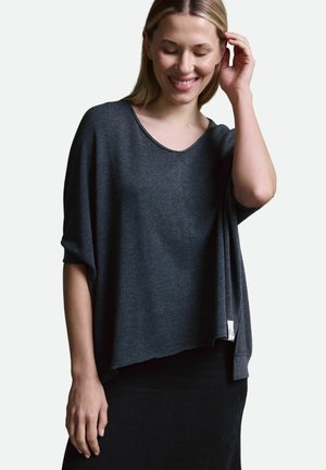 Lächelnde Frau mit blonden Haaren, die ein lockeres, dunkelgraues Kurzarmoberteil und einen schwarzen Rock trägt, während sie mit einer Hand durch ihr Haar fährt.