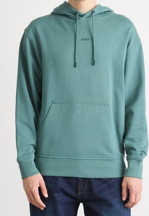 Homme portant un sweat à capuche bleu sarcelle avec poche avant et logo « BOSS », associé à un jean bleu foncé.