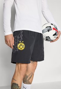 Pantalones cortos deportivos negros con un patrón texturizado y un logo amarillo de Borussia Dortmund, combinados con una camiseta blanca de manga larga y un balón de fútbol.