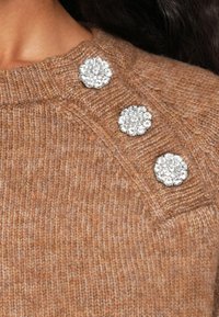 Brązowy sweter dzianinowy o teksturowym wykończeniu, z trzema srebrnymi ozdobami w kształcie klejnotów umieszczonymi na ramieniu.