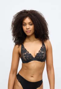 Melna mežģīņu bralette ar ziedu rakstu, dziļu dekoltē un caurspīdīgu audumu. Kombinēta ar atbilstošām melnām apakšveļas biksītēm.
