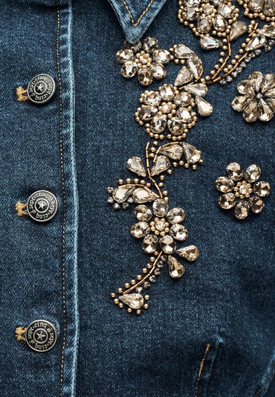 Ύφασμα από τζιν με ασημένια κουμπιά και περίτεχνη κεντητική με λουλούδια από χρυσά και διαφανή πολύτιμους λίθους και χάντρες στο μπροστινό μέρος ενός μπουφάν.