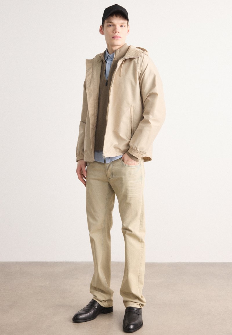 Veste à capuche beige sur un pull marron, une chemise bleu clair et un pantalon beige ; chaussures noires ; style décontracté ; tissu lisse et coupe relax. 