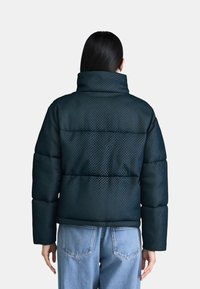 Giacca piumino verde scuro con colletto alto, caratterizzata da un motivo geometrico, texture trapuntata e maniche ampie. Abbinata a jeans azzurri chiari.