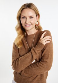 Brauner Strickpullover mit Rundhalsausschnitt, über einem weißen Oberteil getragen. Ergänzt mit großen runden Ohrringen und einer langen silbernen Halskette.