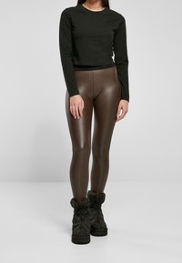 Nainen, jolla on päällään musta pitkähihaisena crop-top, kiiltävät ruskeat legginsit ja mustat eristävät nauhakengät, seisoo tasaisella taustalla.