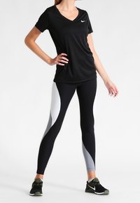 Maglietta nera a V con un piccolo logo bianco, abbinata a leggings neri con pannelli grigi. Il modello indossa scarpe da ginnastica nere.