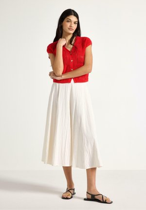 Donna in piedi con le braccia incrociate, indossa una maglia rossa a maniche corte con bottoni, una gonna midi bianca e sandali neri su sfondo bianco.