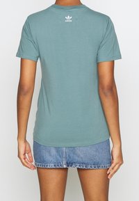 Tealfärgad bomullströja med rund hals och korta ärmar, med en vit Adidas-logotyp tryckt på den övre ryggen. Bärs med en denimkjol.