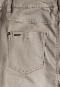 Pantalons beiges avec une poche arrière unique dotée d'un bouton en métal et d'une petite étiquette de marque métallique, en tissu lisse avec une légère brillance.