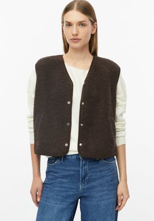 Frau trägt eine dunkelbraune Fleeceweste mit Druckknöpfen über einem weißen Langarmshirt und blauen Jeans, steht vor einem einfarbigen Hintergrund.