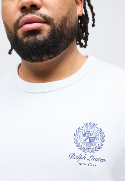 Άνδρας με γενειάδα που φοράει λευκό μπλουζάκι με στρογγυλή λαιμόκοψη και μπλε λογότυπο Polo Ralph Lauren New York στο στήθος.