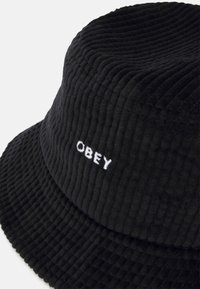 Crni rebrasti šešir u obliku kante izrađen od kordura tkanine, sa izvezenim bijelim logom "OBEY" sprijeda. Okrogli, strukturirani dizajn s mekom teksturom.