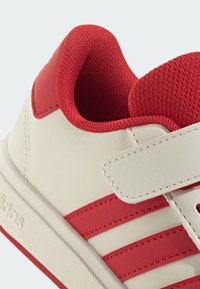 Bílé tenisky s červenými akcenty. Mají hladký syntetický svršek, polstrovaný límec, suchý zip a tři červené pruhy na boku.