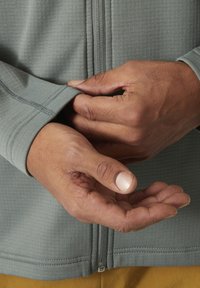 Chaqueta ligera de manga larga con cremallera en tejido gris con un patrón texturizado. Primer plano de manos ajustando el puño y el detalle de la cremallera.