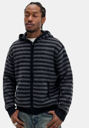 Homme aux cheveux tressés portant un sweat à capuche zippé rayé noir et gris et un jean bleu, debout, les mains dans les poches, devant un fond uni.