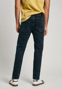 Dark blue denim jeans with a slim fit, featuring two back pockets and a mid-rise waist. Paired with white sneakers and a yellow shirt.