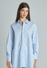 Camicia a righe azzurro chiaro con maniche lunghe, design con bottoni e decorata con dettagli argentati. Presenta un colletto classico e polsini.