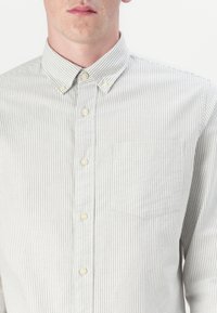 Camisa de botones fabricada en un tejido ligero, con rayas en gris claro y blanco, un cuello y un bolsillo en el pecho. Los botones son de color claro.