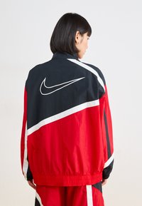 Nike vetrovka z barvno kombinacijo črne, rdeče in bele, z velikim belim logotipom na hrbtu. Lahka najlonska tkanina.