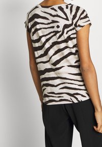 Person klädd i en kortärmad tröja med zebrarandigt mönster i vitt och svart samt svarta byxor, visad bakifrån mot en neutral bakgrund.