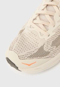 Μπεζ αθλ ητικό παπούτσι με υφασμένη υφή στο επάνω μέρος, διαθέτει ανοιχτόχρωκα κορδόνια, ευέλικτη σόλα και πορτοκαλί λεπτομέρεια κοντά στη μύτη.