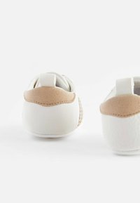 Zapatos de bebé de cuero blanco con acentos de gamuza beige, forma redondeada y una lengüeta en el talón en bucle. Textura suave y detalles de costura.
