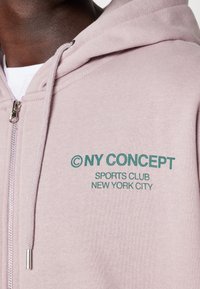 Sudadera con capucha y cremallera en rosa claro, hecha de mezcla de algodón, con texto verde que dice "© NY CONCEPT SPORTS CLUB NEW YORK CITY."