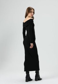 Schwarzes schulterfreies Kleid mit langen Ärmeln und Raffung, taillierter Silhouette und bodenlangem Design, kombiniert mit schwarzen kniehohen Stiefeln.