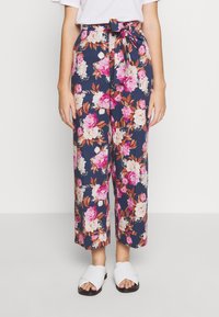 Nainen, joka on pukeutunut tummansinisiin leveisiin housuihin, joissa on vaaleanpunaisia ja valkoisia kukkakuoseja, vyötärölle solmittu vyö ja valkoiset avokaulatossut.