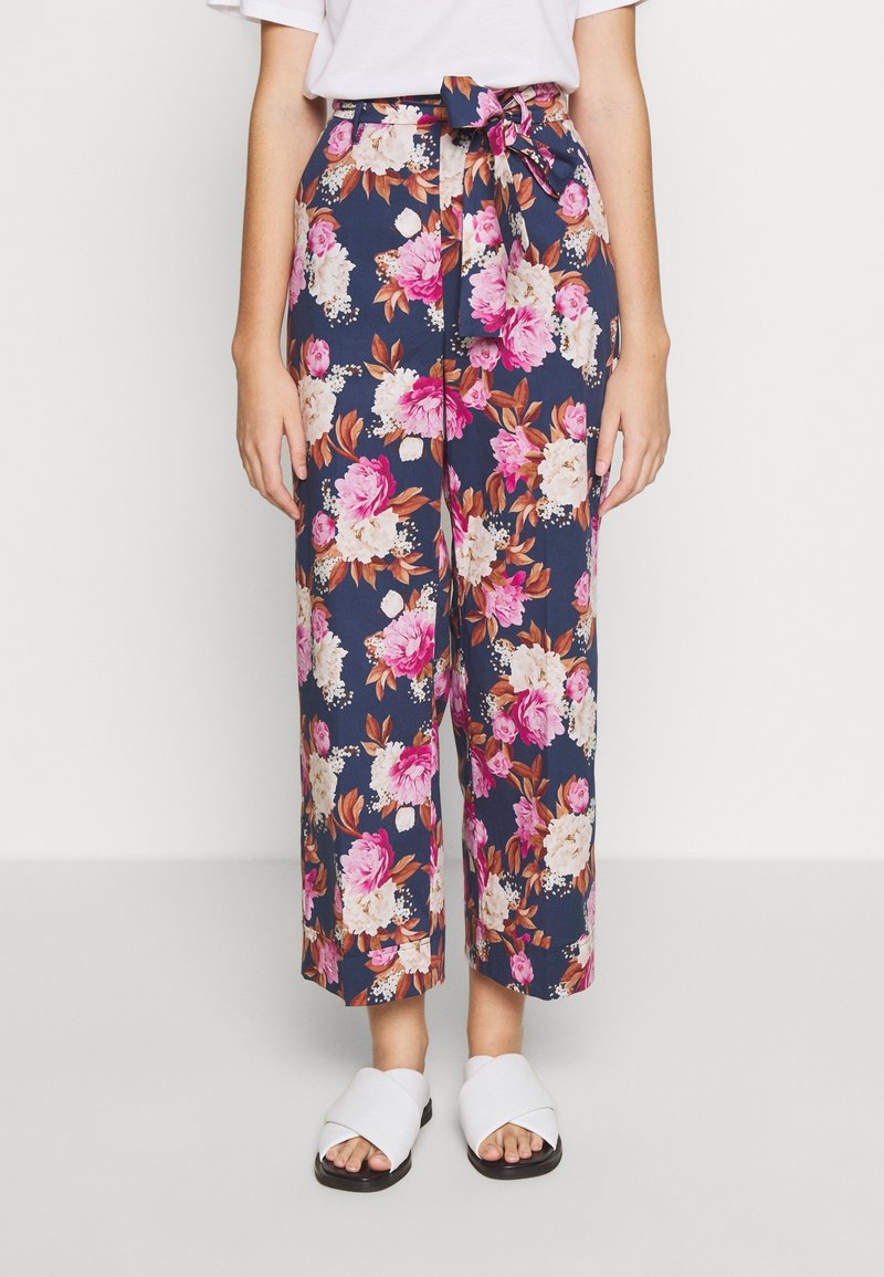 Nainen, joka on pukeutunut tummansinisiin leveisiin housuihin, joissa on vaaleanpunaisia ja valkoisia kukkakuoseja, vyötärölle solmittu vyö ja valkoiset avokaulatossut.