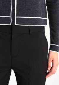 Pull cropped gris foncé avec rayures blanches, doté de deux poches avant et d'une fermeture à boutons, associé à un pantalon noir ajusté.